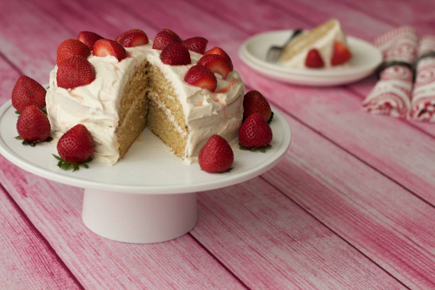 Vegan vanilla cake with one slice removed on a white cake stand. 