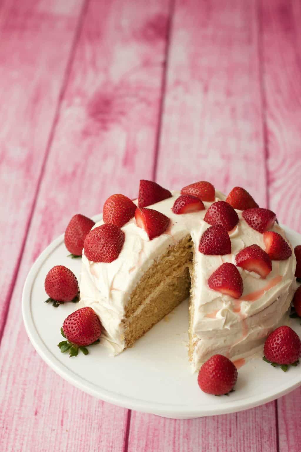 Vegan vanilla cake topped with fresh strawberries on a white cake stand. 