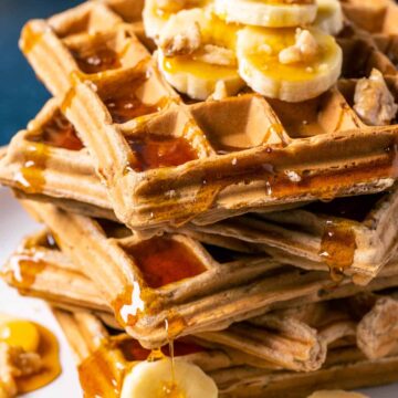 Stack of vegan banana waffles topped with sliced banana, walnuts and syrup on a white plate.