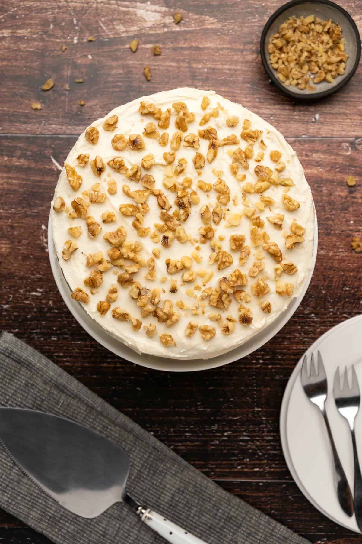 Vegan carrot cake topped with crushed walnuts on a white cake stand.