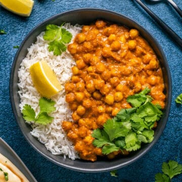 Vegan chana masala in a black bowl with rice, cilantro and lemon.