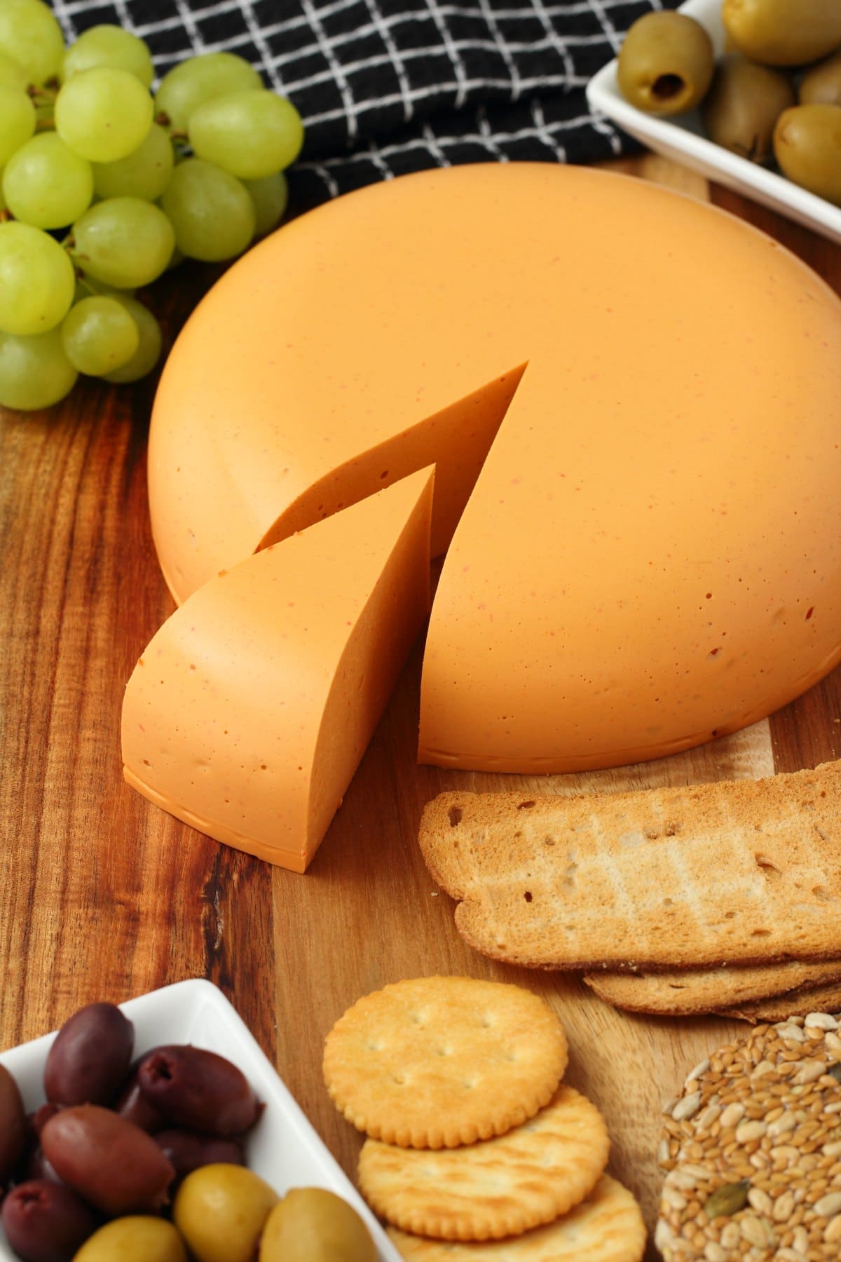 Vegan cheddar cheese round on a cheese board with crackers olives and grapes. 