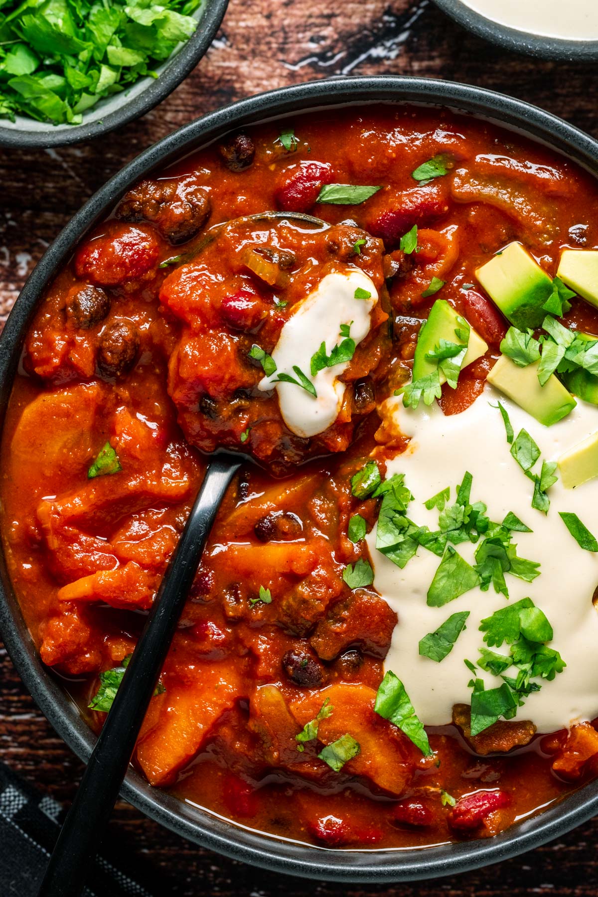 Vegan chili with chopped cilantro, cashew cream and diced avocado in a black bowl with a spoon.