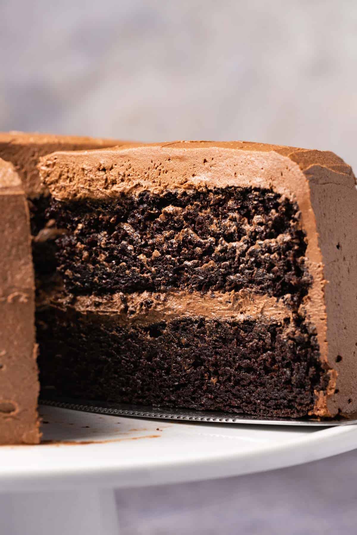 Vegan chocolate cake on a white cake stand with one slice cut and ready to serve. 