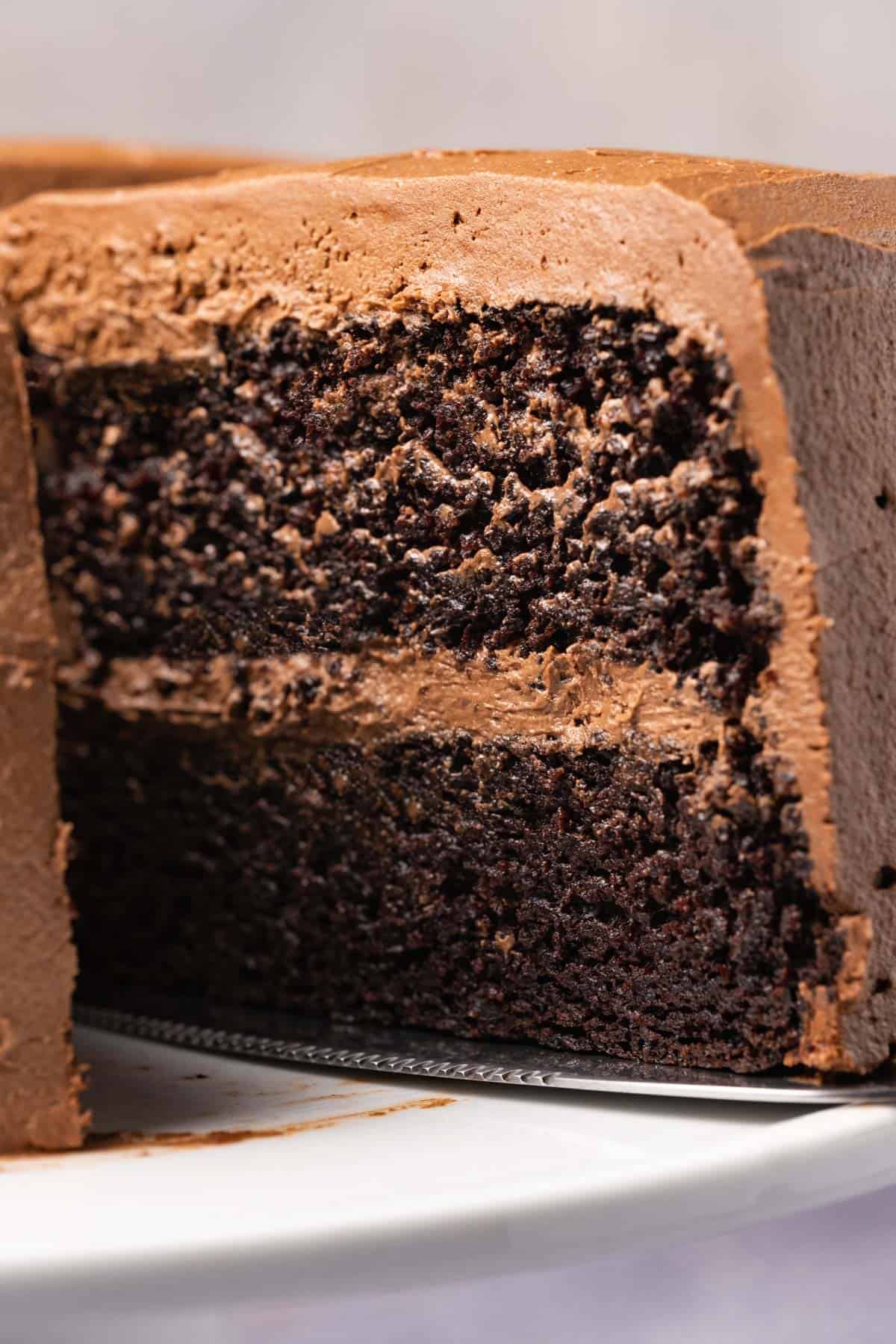 Vegan chocolate cake on a white cake stand with one slice cut and ready to serve.