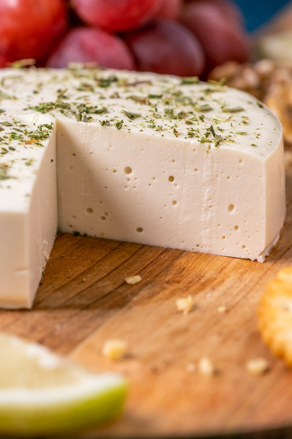 Round of vegan goat cheese with slices removed on a wooden board with crackers, nuts and grapes.