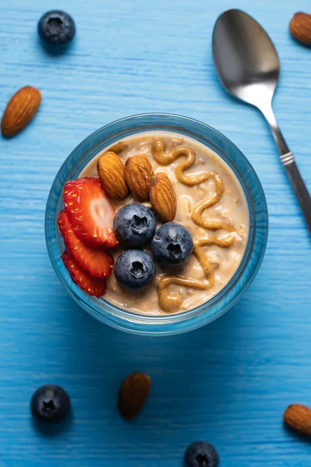 Vegan overnight oats topped with drizzled almond butter, blueberries and strawberries in a glass jar. 