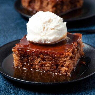 Slice of vegan sticky toffee pudding topped with ice cream on a black plate.