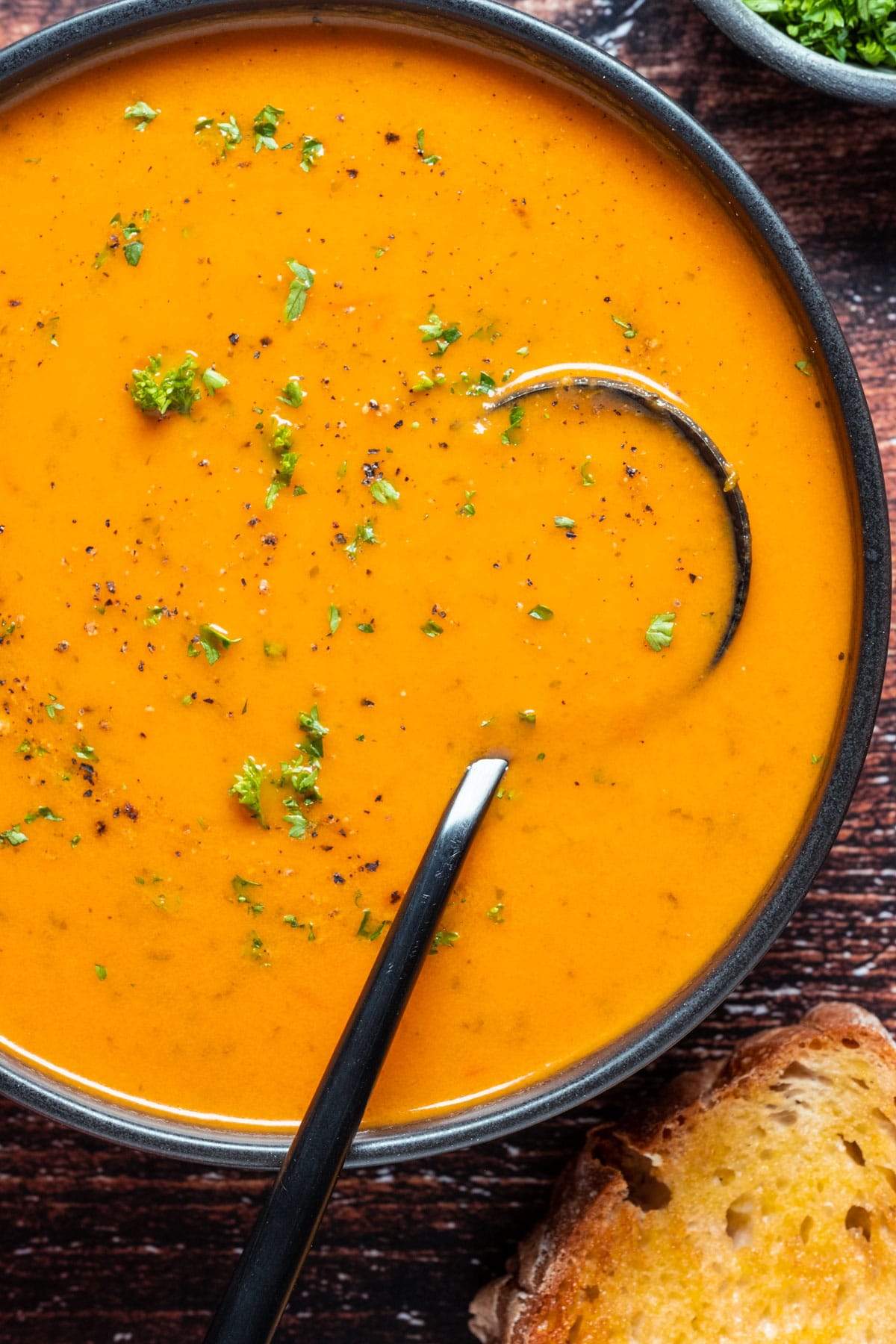 Vegan tomato soup topped with fresh chopped parsley in a black bowl with a spoon.