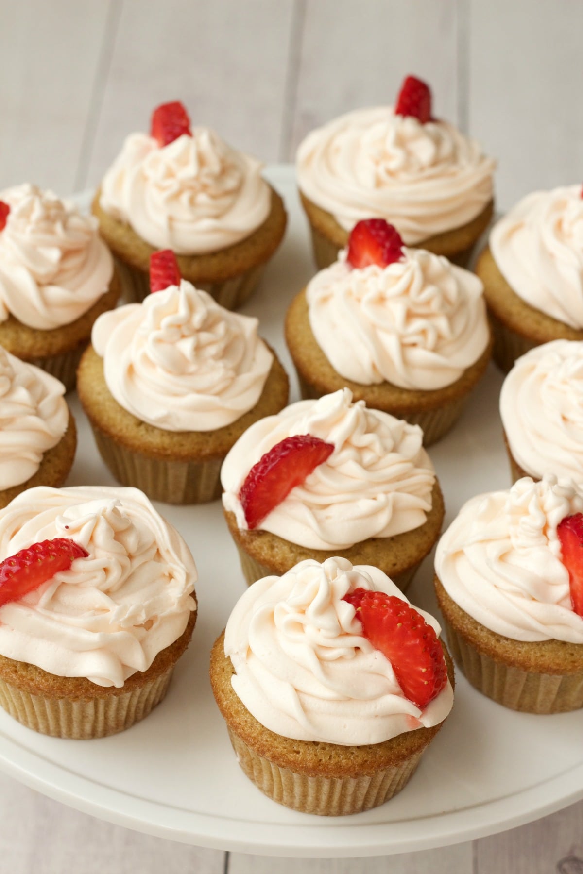 Vegan vanilla cupcakes with strawberry vanilla frosting and fresh strawberry pieces on a white cake stand.