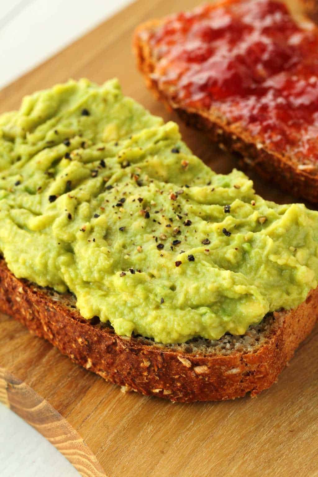 Two slices of whole wheat bread on a wooden board, one topped with avocado and the other topped with strawberry jam. 